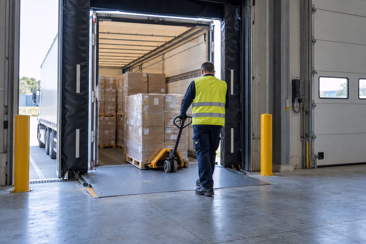 Worker moving a pallet of boxes from a truck at a warehouse loading dock with a pallet jack