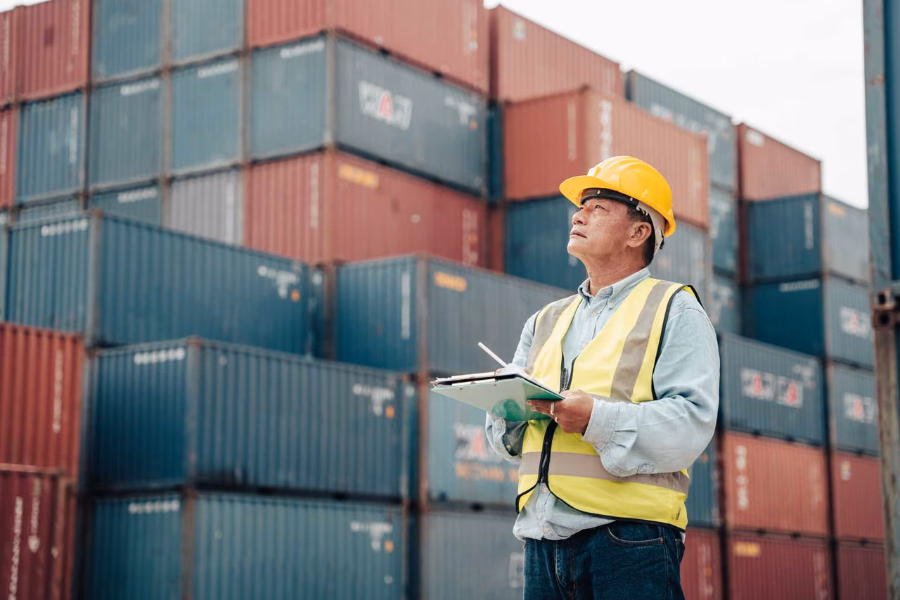 Inspector in a yellow hard hat and high-visibility vest holding a clipboard between stacked shipping containers